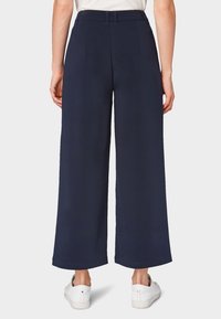 Osoba w granatowych, szerokich, krótko obciętych spodniach i białych trampkach, stojąca plecami na tle jednolitego tła.