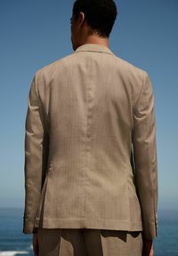 Giacca da suit beige chiaro con finitura testurizzata, con una fessura singola sul retro e sottili righe verticali, inquadrata contro un cielo azzurro chiaro.