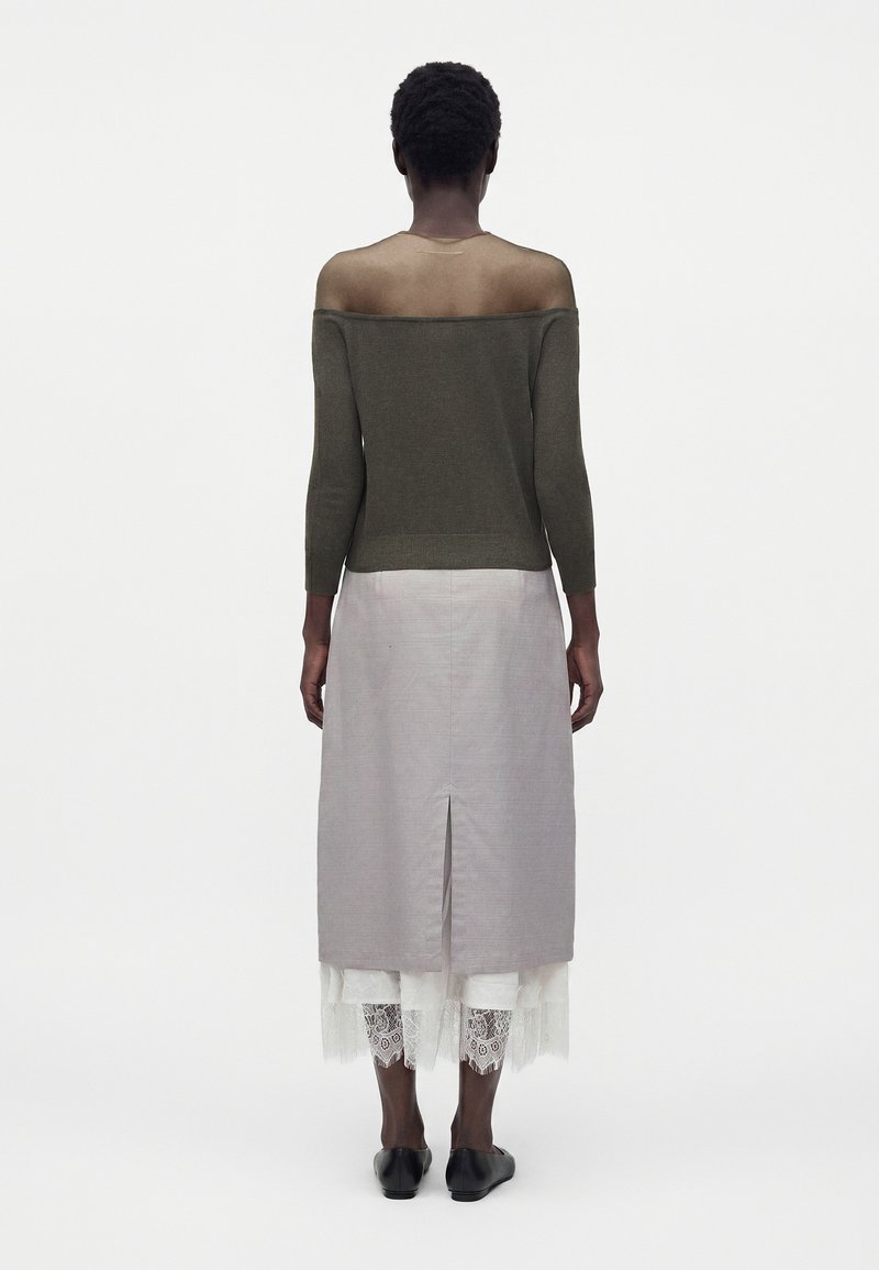 Woman wearing an off-the-shoulder olive green sweater, light gray skirt with back slit, lace-trimmed white underlayer, and black flats, standing.