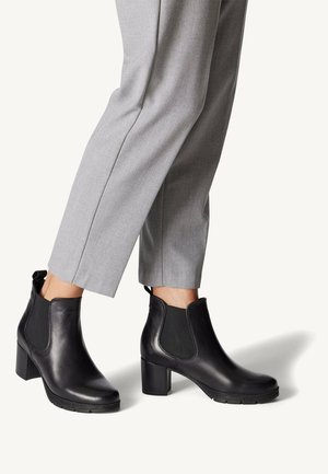 Bottines noires en cuir avec un talon bloc épais, dotées de panneaux élastiques sur les côtés et d'une languette pour un enfilage facile, associées à un pantalon gris ajusté.