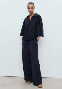 Černý oversized top s límečkem, kombinovaný s volnými černými kalhotami vyrobenými z měkké látky. Minimalistický design a uvolněná silueta.