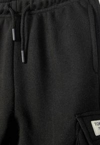 Gros plan sur un pantalon de survêtement noir avec une taille à cordon et une poche latérale ornée d'un patch rectangulaire blanc avec du texte.