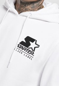 Felpa bianca con logo "Starter Black Label" nero sul petto, indossata da una persona con tatuaggio sul collo e collana d'oro.