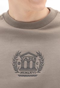 Sudadera beige con cuello redondo acanalado, que presenta un logo oscuro bordado de un edificio y ramas de laurel, con texto detallado debajo.