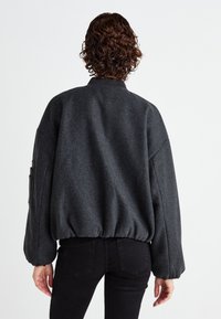 Siva oversized bomber jakna s rebrastim pojasom i manžetama, koja se odlikuje visokim ovratnikom i blago teksturiranim materijalom.