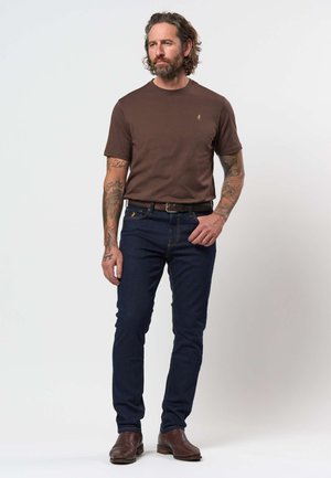 Moški s tetovažami, ki nosi rjavo kratko srajco, temno modre kavbojke, rjav pas in rjave škornje, stoji na belem ozadju.