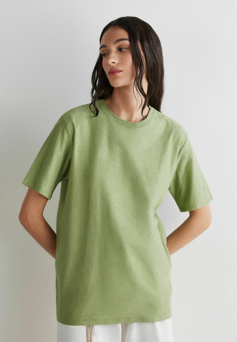 Vrouw met lang donker haar, gekleed in een losse lichtgroene korte mouw blouse, staand met handen achter de rug tegen een effen witte muur.