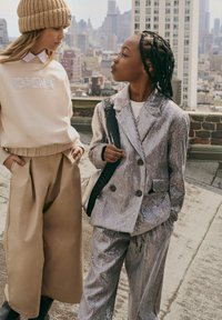 Dwoje dzieci na dachu, jedno w beżowym swetrze i szerokich spodniach, drugie w srebrnym garniturze z cekinami z warkoczem i plecakiem.