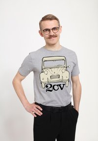 Šedé bavlněné tričko s grafikou klasického auta a textem "2cv" v tučném černém písmu. Modelka nosí černé kalhoty a pózuje s jednou rukou na boku.