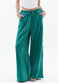 Wijdvallende broeken van glanzende teal stof met een elastische taille, trekkoordsluiting en zijzakken. Soepele textuur met een ontspannen pasvorm.
