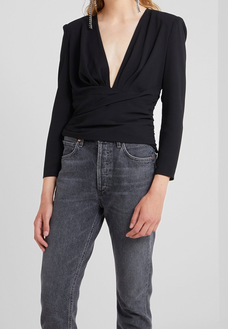Blouse noire à manches longues avec un décolleté en V profond et un détail de taille froncée, associée à un jean gris délavé taille haute. Texture lisse, design ajusté.