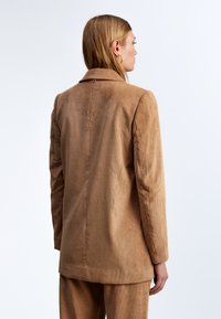 Blazer in corduroy marrone con colletto a revers, texture a coste verticali e taglio dritto. La vista posteriore mostra i dettagli delle spalle e le cuciture.