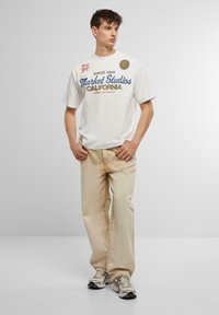 Balta kokvilnas T-kreklu ar zilu un sarkanu tekstu, kombinēts ar gaiši beige džinsiem. Modelis valkā sudraba sporta apavus. Vienkārša fona.