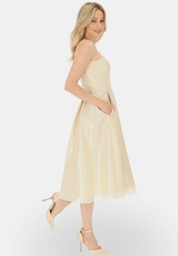 Jasno beżowa sukienka w kształcie litery A z ramiączkami, teksturowany materiał, ściągnięta w talii i kieszeniami. Modelka w nagich szpilkach z paskiem na kostce.