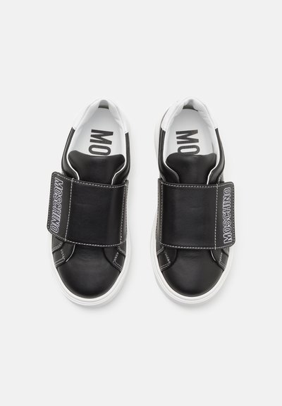 Zapatos de cuero negro sin cordones con una suela blanca y una correa de velcro, con costuras plateadas y la marca "Moschino" en la correa.