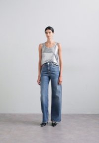 Top plateado sin mangas, jeans anchos de mezclilla azul claro de cintura alta, zapatos negros de punta afilada, fondo liso.