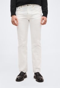 Άνδρας που φοράει εκρού τζιν με ίσια γραμμή, μαύρα μοκασίνια και σκούρο πουλόβερ με μακριά μανίκια, στέκεται μπροστά σε απλό φόντο.