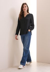 Camicia nera a maniche lunghe con bottoni, abbinata a jeans blu a gamba dritta e sneakers bianche, su uno sfondo chiaro.