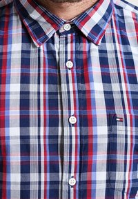 Närbild på en person som bär en rutig skjorta i röd, blå och vit färg med krage och bröstficka.