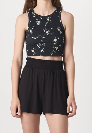 Kobieta ubrana w czarny, bezrękawnik w kwiaty i czarne, krótkie spodenki z wysokim stanem na jednolitym tle.