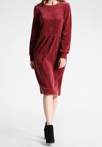 Burgundowa sukienka z weluru z długimi rękawami, okrągłym dekoltem i zbieraną talią. Długość do połowy łydki, o gładkiej strukturze i subtelnym blasku.