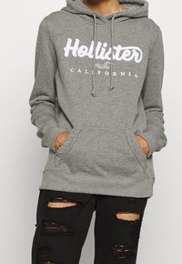 Felpa grigia con logo bianco "Hollister California". Presenta un cappuccio con cordoncino, tasche frontali e polsini e orlo a coste. Indossata con jeans neri strappati.