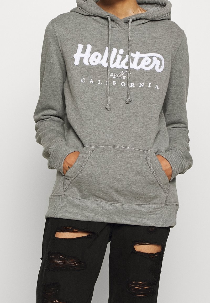 Felpa grigia con logo bianco "Hollister California". Presenta un cappuccio con cordoncino, tasche frontali e polsini e orlo a coste. Indossata con jeans neri strappati.