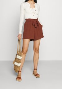 Un haut en tricot léger couleur crème à manches longues avec des boutons, associé à un short taille haute couleur rouille et des sandales à lanières marron. Tient un sac en osier.