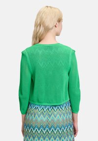 Grüne gestrickte Strickjacke mit Dreiviertelärmeln, die ein lockeres, strukturiertes Gewebe aufweist. Der Rücken ist kurz und betont einen Rock mit Zickzackmuster.