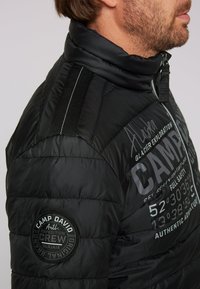 Giacca a vento nera con texture a trapuntatura, caratterizzata da grande testo stampato e un patch rotondo sul braccio sinistro. Design con zipper e collettore alto.