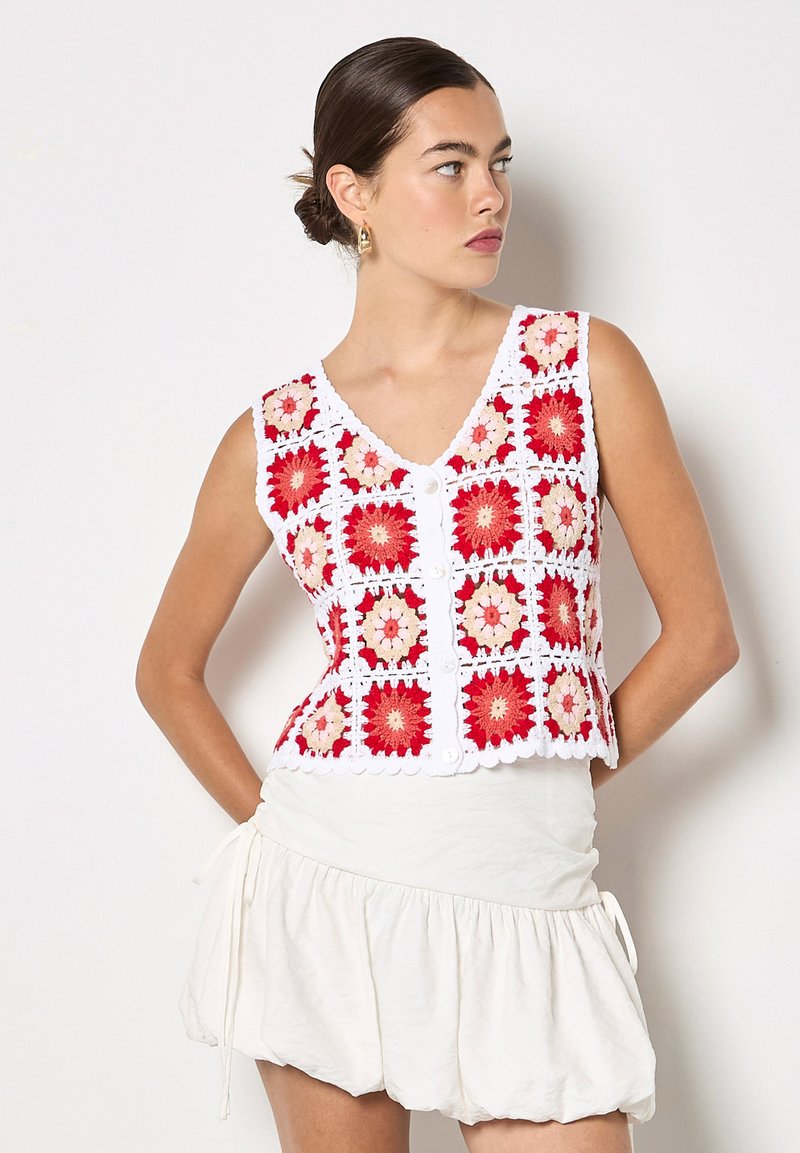 Femme aux cheveux noirs attachés en chignon, portant un gilet blanc en crochet sans manches orné de carrés floraux rouges et roses, associé à une jupe blanche à volants.