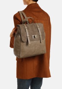 Grande zaino in pelle grigia con chiusura a pattina, due spallacci e dettagli metallici, indossato sopra una giacca in suede marrone.