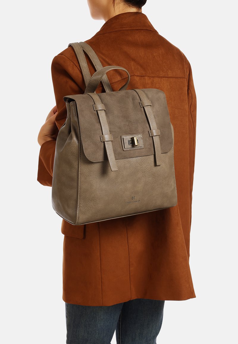 Grande zaino in pelle grigia con chiusura a pattina, due spallacci e dettagli metallici, indossato sopra una giacca in suede marrone.