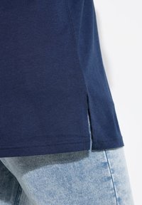 T-shirt blu navy con una texture liscia, caratterizzato da spacchi laterali all'orlo. Abbinato a jeans chiari, che mostrano un design aderente.