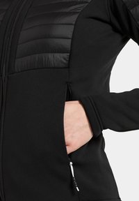 Mâna unei persoane în buzunarul lateral cu fermoar al unei jachete negre, cu secțiune superioară matlasată și mâneci din material textil.