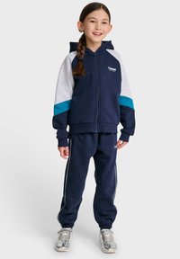 Felpa con cappuccio blu navy con dettagli grigi e turchesi, chiusura a zip, pantaloni della tuta abbinati con strisce laterali bianche e sneakers metalliche.