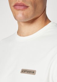 Tricou alb cu guler rotund, având o etichetă rectangulară bej cu textul "ICEPEAK", textură netedă și mâneci scurte.