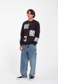 Suéter oscuro con patrones geométricos blancos, combinado con pantalones vaqueros de pierna ancha en azul claro. El conjunto tiene un ajuste casual y relajado.