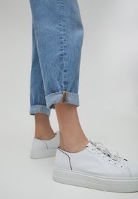 Jeans de color azul claro con el dobladillo desflecado, combinados con zapatillas blancas que tienen una suela texturizada y acentos en negro.