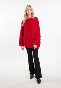 Roter gestrickter Pullover mit strukturierten, gepufften Ärmeln und geripptem Saum, kombiniert mit schwarzen ausgestellten Hosen und spitzen schwarzen Stiefeln.