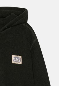 Svart fleece hoodie med en framficka, utrustad med en sydd etikett med en blå bergsgrafik och krämfärgad kant. Texturerat tyg och huva.