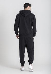 Felpa nera con cappuccio e pantaloni jogger realizzati in tessuto morbido, caratterizzati da una vestibilità rilassata, polsini a costine e cappuccio con cordoncino. Sneakers bianche completano l'outfit.