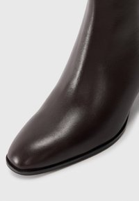 Botte de cheville en cuir marron avec un bout pointu, texture lisse et un talon fin. Design minimaliste avec des détails de couture subtils.