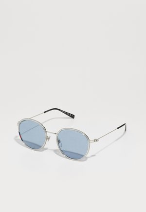 Lunettes de soleil ovales avec des verres bleus, monture en métal argenté fine et embouts de branches noirs. Présente un logo et des accents rouges sur le verre gauche.