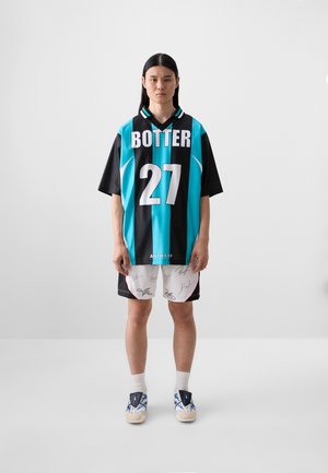 Tricou oversized negru și teal cu textul "BOTTER" și numărul "27", asortat cu pantaloni scurți albi cu semnături. Tenisi albi completează ținuta.