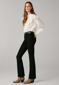 Vaqueros negros acampanados combinados con una blusa blanca con mangas con volante y un acabado de encaje. La modelo lleva botas de tacón de color marrón claro.