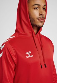 Rote Kapuzenjacke mit einer Fronttasche, Kordelzugkapuze, weißem Logo und seitlichen Streifen. Hergestellt aus glattem Stoff mit einer taillierten Passform.