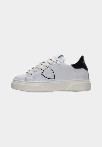 Baskets basses blanches avec lacets beige, semelle épaisse, languette de talon noire, et patch logo bordé de noir sur le côté.