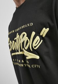 Sort t-shirt med fremtrædende gult grafisk tekst, blød tekstur og urban brandlogo-design, vinkelret for at vise detaljer og pasform.