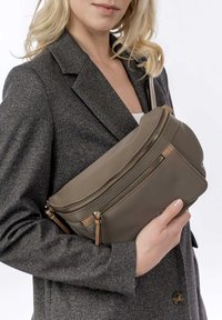 Brązowa skórzana torebka typu crossbody z dwoma zamykanymi na zamek przegrodami, z frędzlami i gładką teksturą. Nosi ją w zestawieniu z szarym żakietem.
